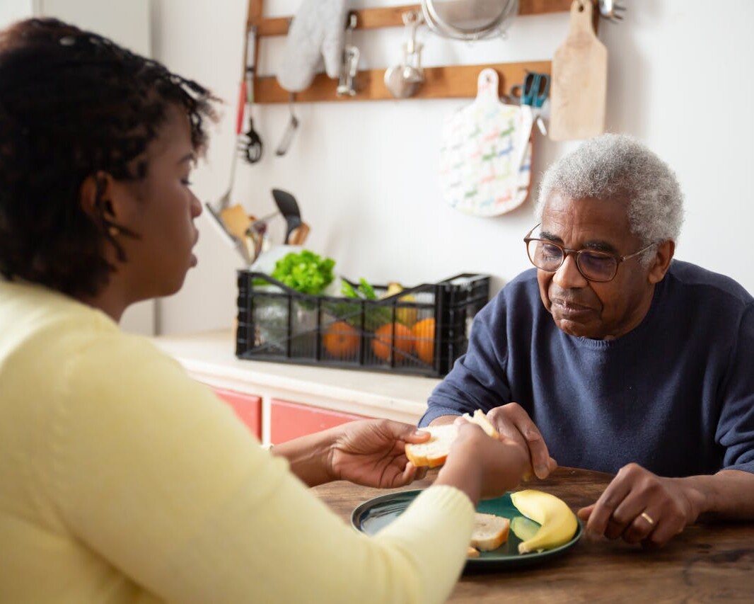 A caregiver and an elderly individual sharing a meal at a wooden table in a cozy home kitchen, representing compassionate in-home care.